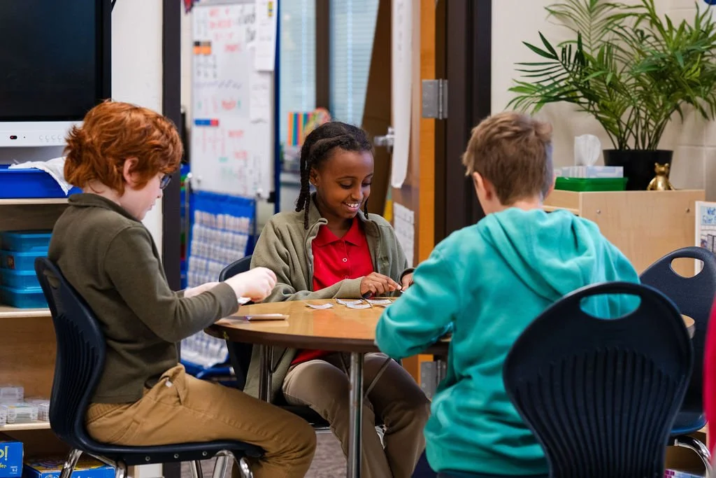 Three children sitting around a table in a classroom, playing a game or working on an activity together.