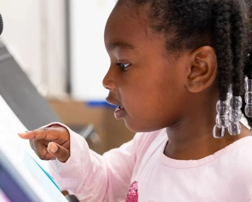 A young girl with dark curly hair and a beaded hair accessory, wearing a light pink shirt, intently looking and touching a screen, possibly a tablet.