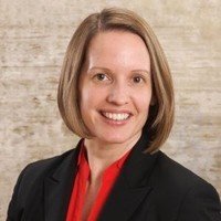 A woman with shoulder-length light brown hair, wearing a black blazer and red blouse, smiling.