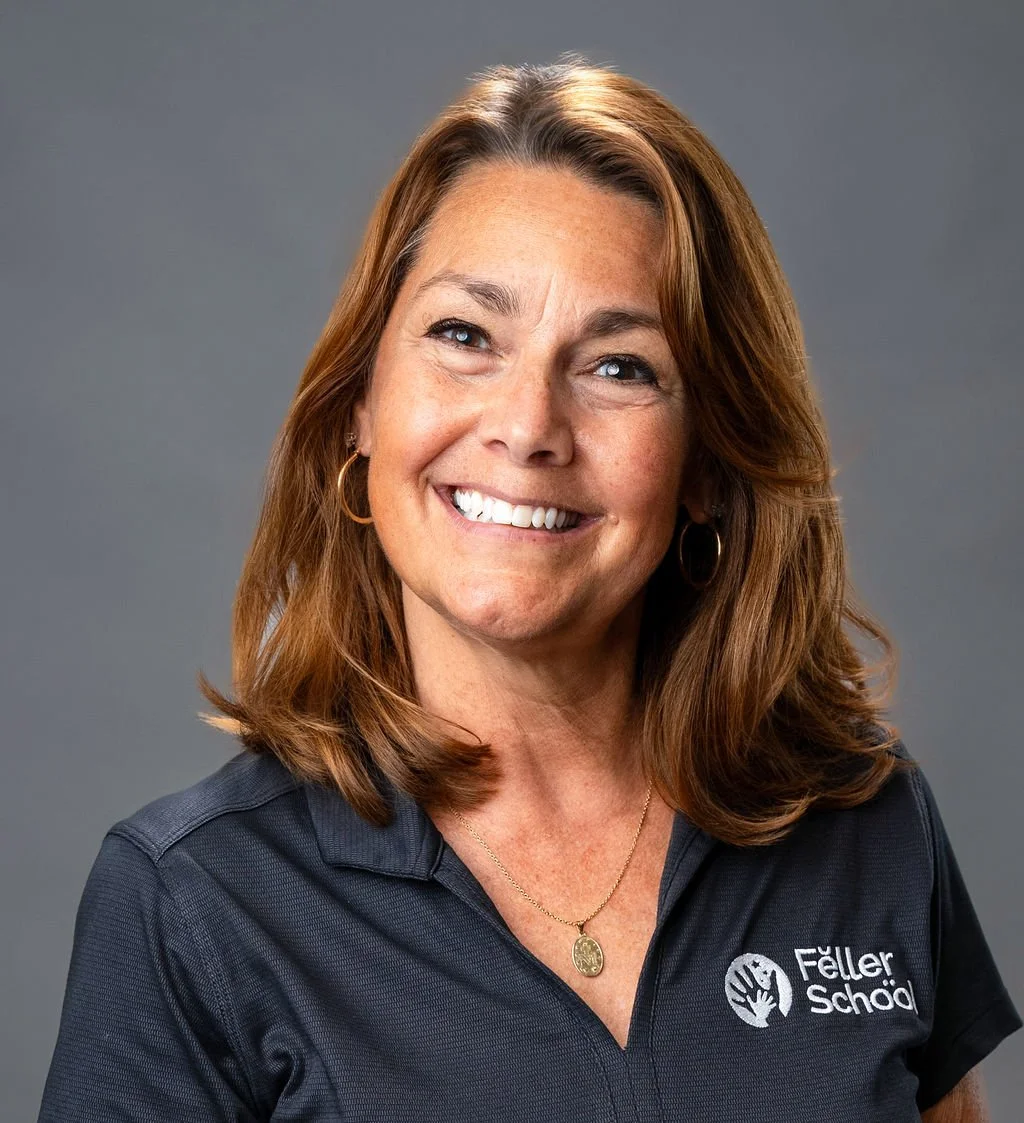 A woman with shoulder-length red hair smiling, wearing a navy blue polo with Feller School logo, gold hoop earrings, and a gold necklace, against a gray background.