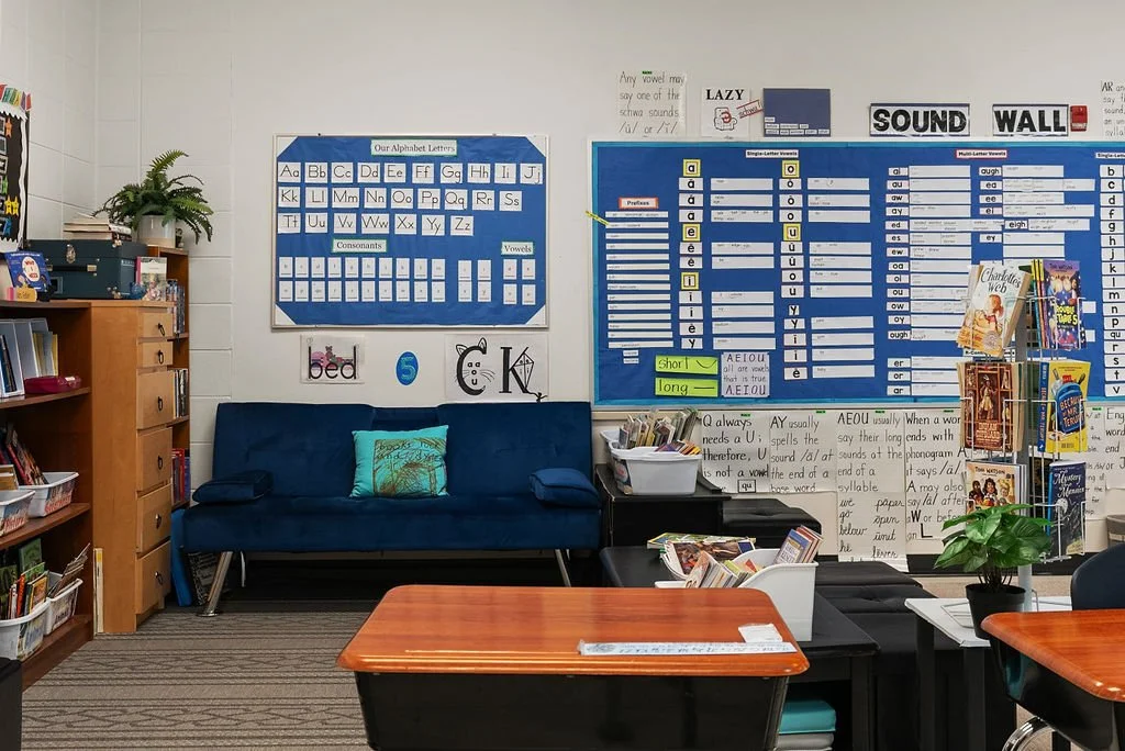 Classroom with educational posters about alphabet letters, sounds, and vowels, a blue couch with pillows, bookshelves filled with books, and tables with educational materials.