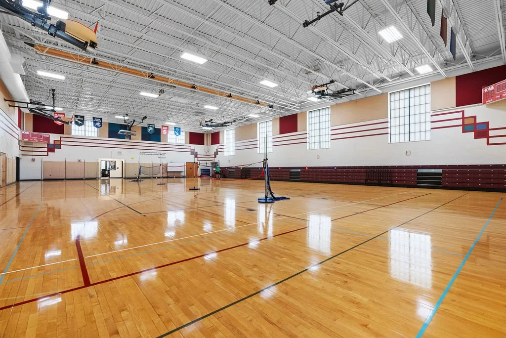 Empty indoor gymnasium with hardwood floor, high ceiling, sports banners, and large windows.