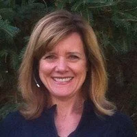 A smiling woman with shoulder-length brown hair standing outdoors in front of greenery.
