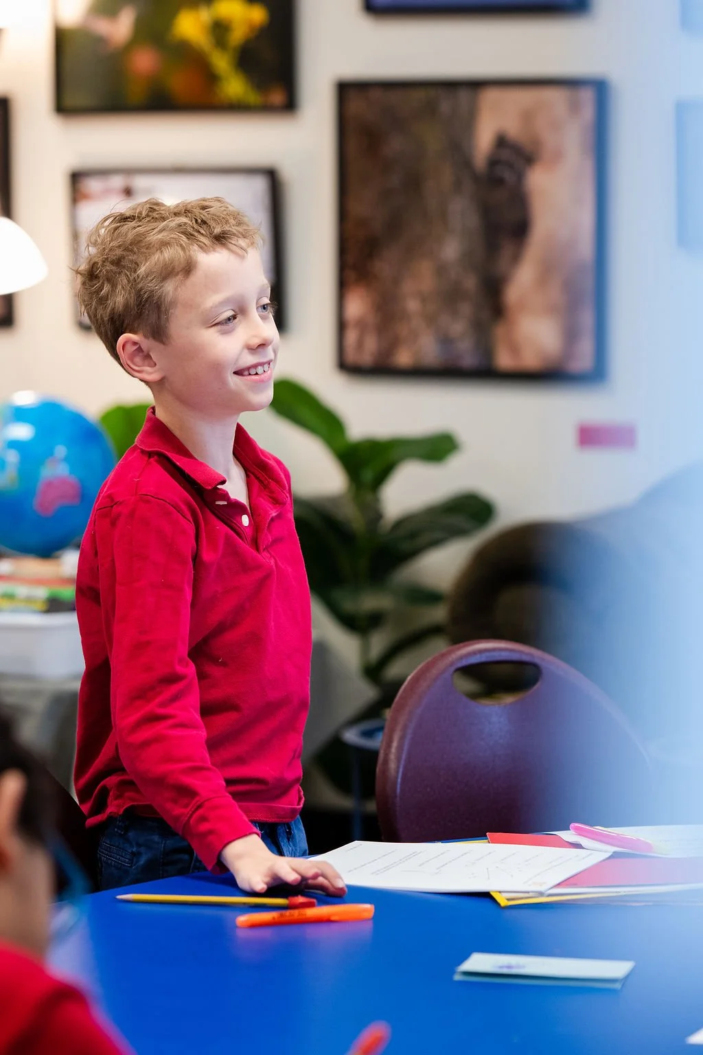 A young boy with curly hair and wearing a red shirt stands at a table with papers and colored markers, smiling in a classroom or art room with framed artwork and a plant in the background.