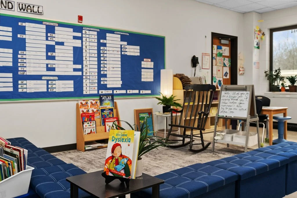 A classroom with a large blue educational wall chart showing phonics and spelling patterns, a whiteboard with handwritten notes, a rocking chair, a small bookshelf filled with children's books, and a cozy seating area with a blue sofa. There are potted plants and artwork on the walls.