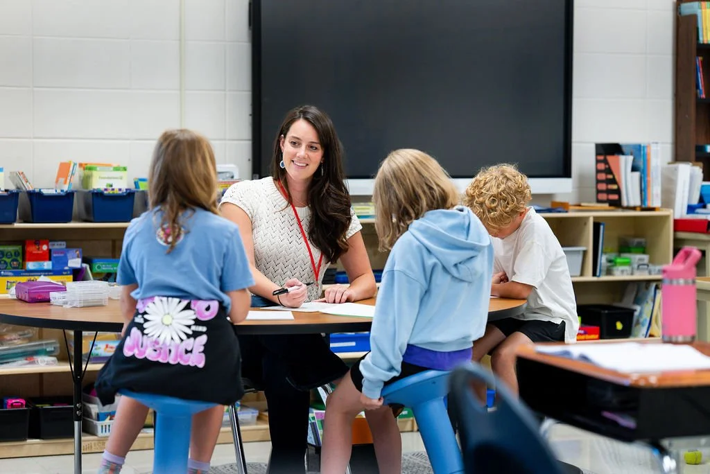 A teacher sitting at a desk smiling and talking to three young children in a classroom, with educational materials and books on shelves in the background.