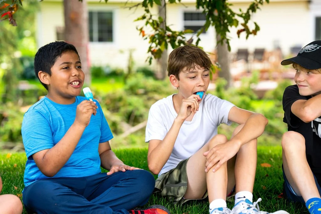 Three kids sitting on grass outdoors, eating popsicles and talking.