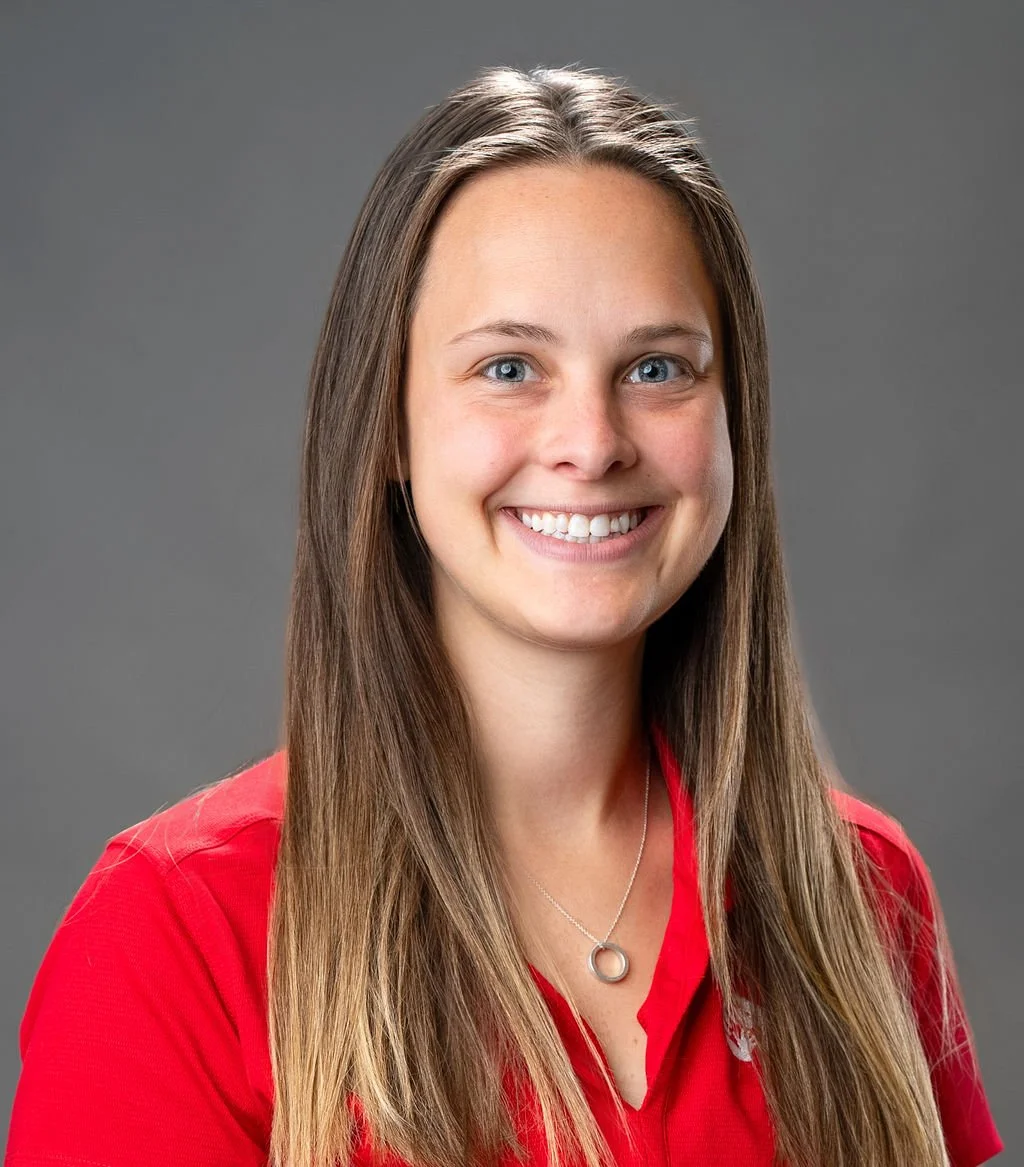 A young woman with long brown hair smiling outdoors, with trees and greenery in the background.