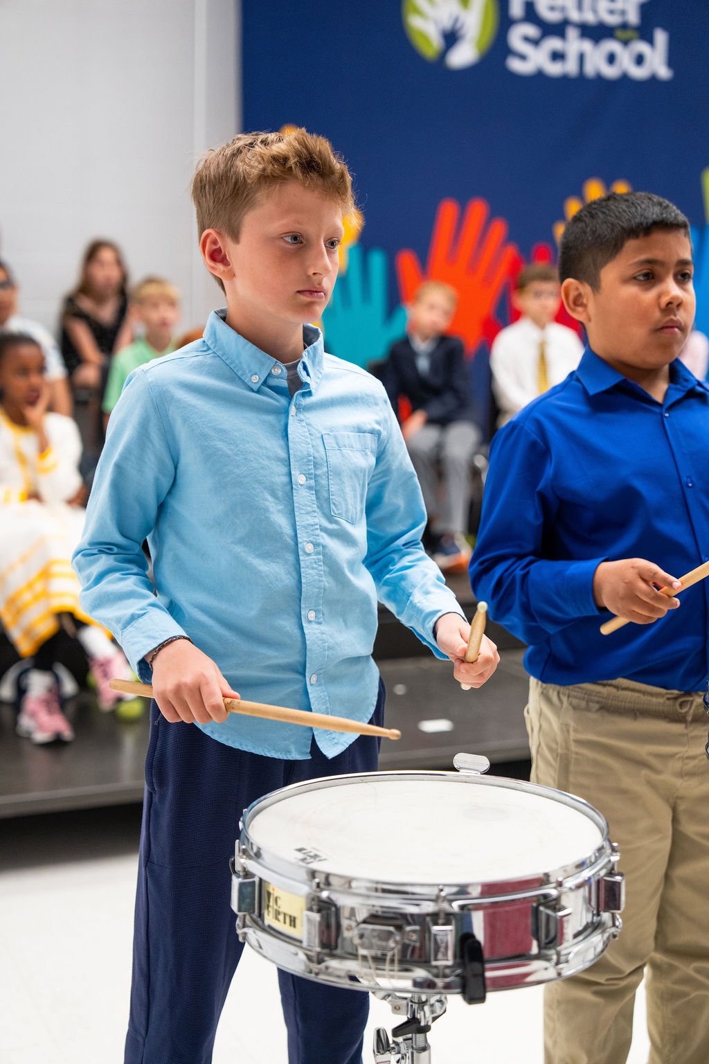 Young boy in a light blue shirt playing a snare drum during a school event, with other children in the background.