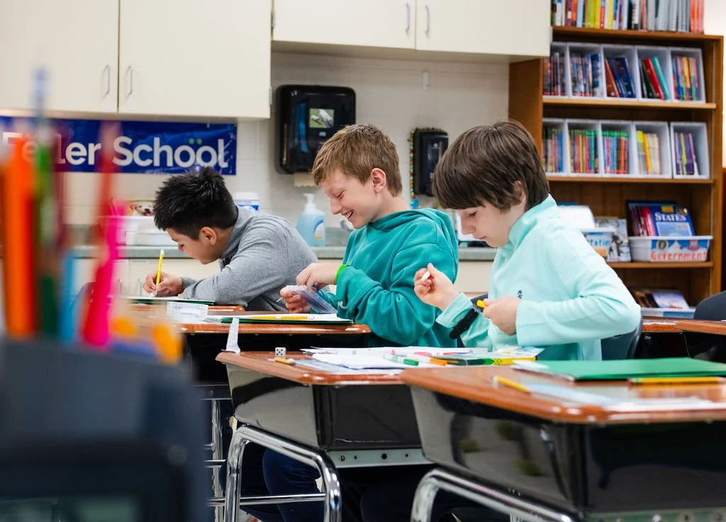 Three middle school students working at desks in a classroom, smiling and concentrating on their schoolwork.