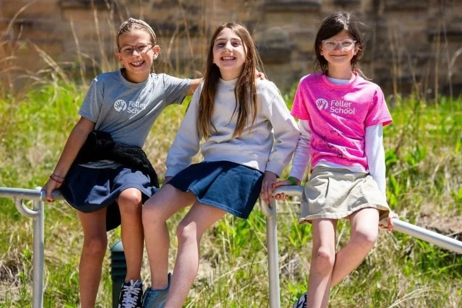 Three young girls sitting on a metal railing outdoors, smiling, with green grass and a wooden fence in the background.