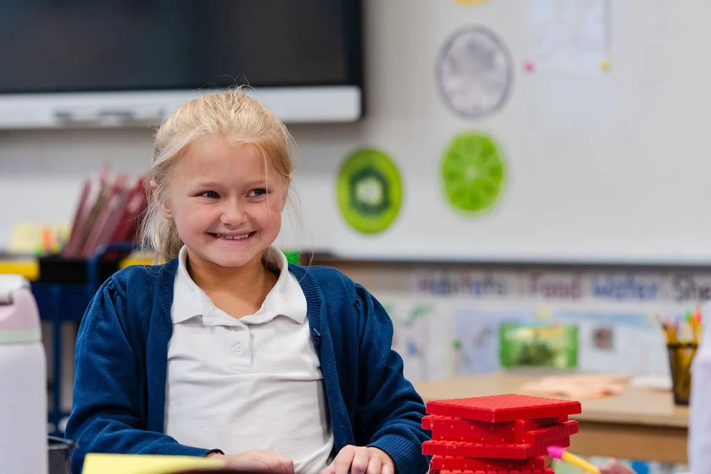 A young girl with blonde hair smiling while sitting at a desk in a classroom, with educational posters and a whiteboard in the background.
