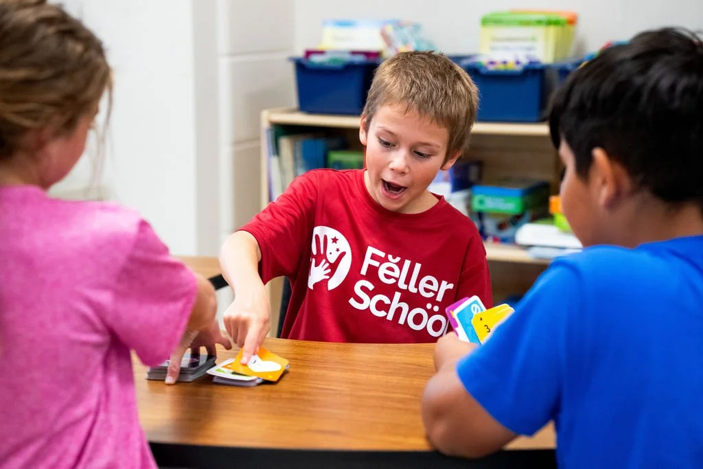 Three children playing a card game at a table in a classroom.