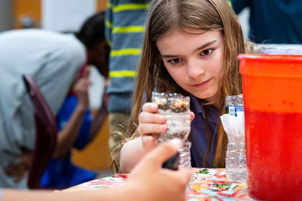 A young girl with long brown hair looking at a plastic bottle filled with rocks and sand on a table, with other children in the background.