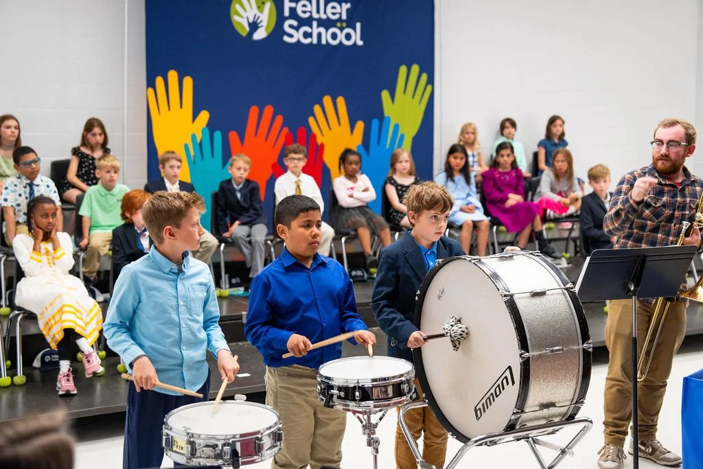 A classroom with children seated on risers, some wearing formal clothes, and others in casual attire, with a large colorful Feller School banner on the wall. Three boys are in front playing drums while a music instructor guides them.