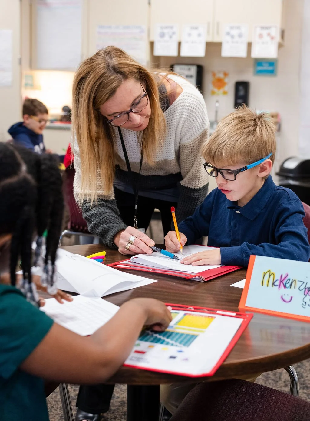 A classroom scene with a female teacher helping a young student with glasses at a table. The teacher is leaning over, pointing at a notebook, while the student writes. Other children are seated at the table, focusing on their work. A colorful sign with the name 'McKenzie' and a smiley face is on the table.