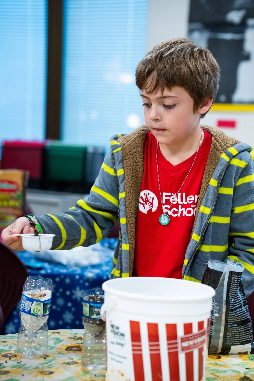 A young boy wearing a red shirt with a logo and a gray hoodie with yellow stripes, standing at a table with popcorn bags and a white bucket, pouring something into a small container in an indoor setting.