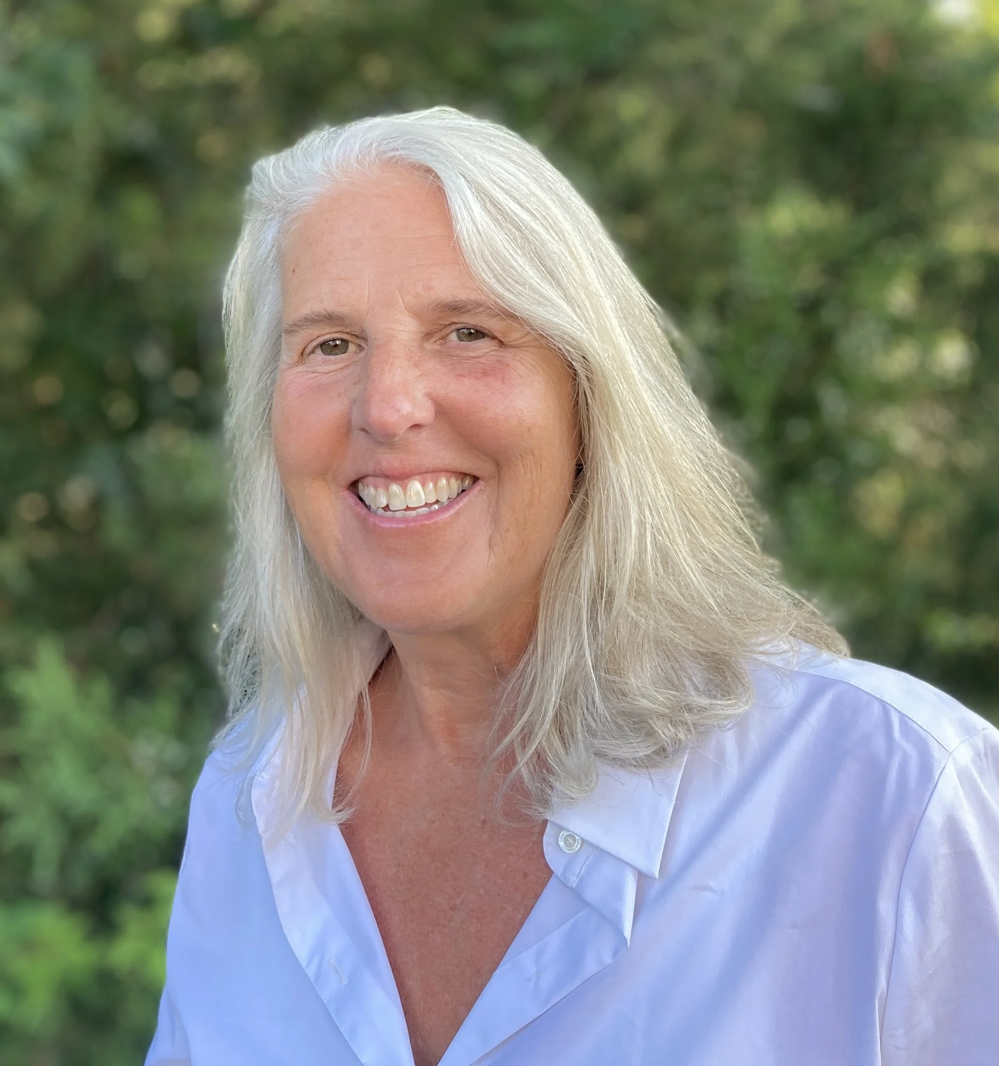 A smiling woman with long, wavy gray hair wearing a light blue shirt outdoors with greenery in the background.