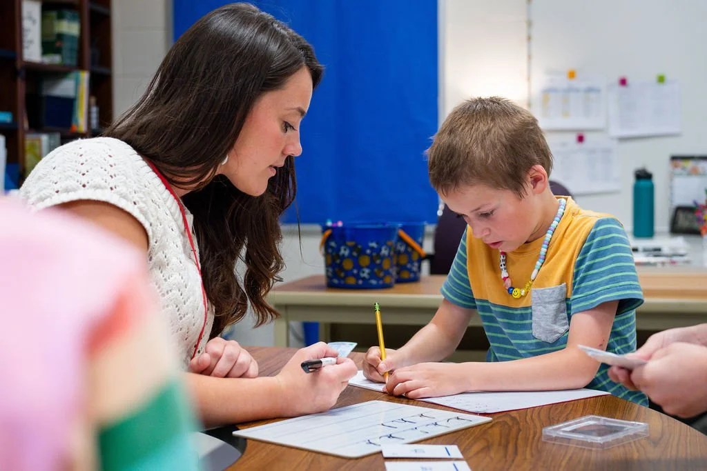 A young woman and boy are sitting at a desk in a classroom, working together on a writing activity, with papers and markers in front of them.