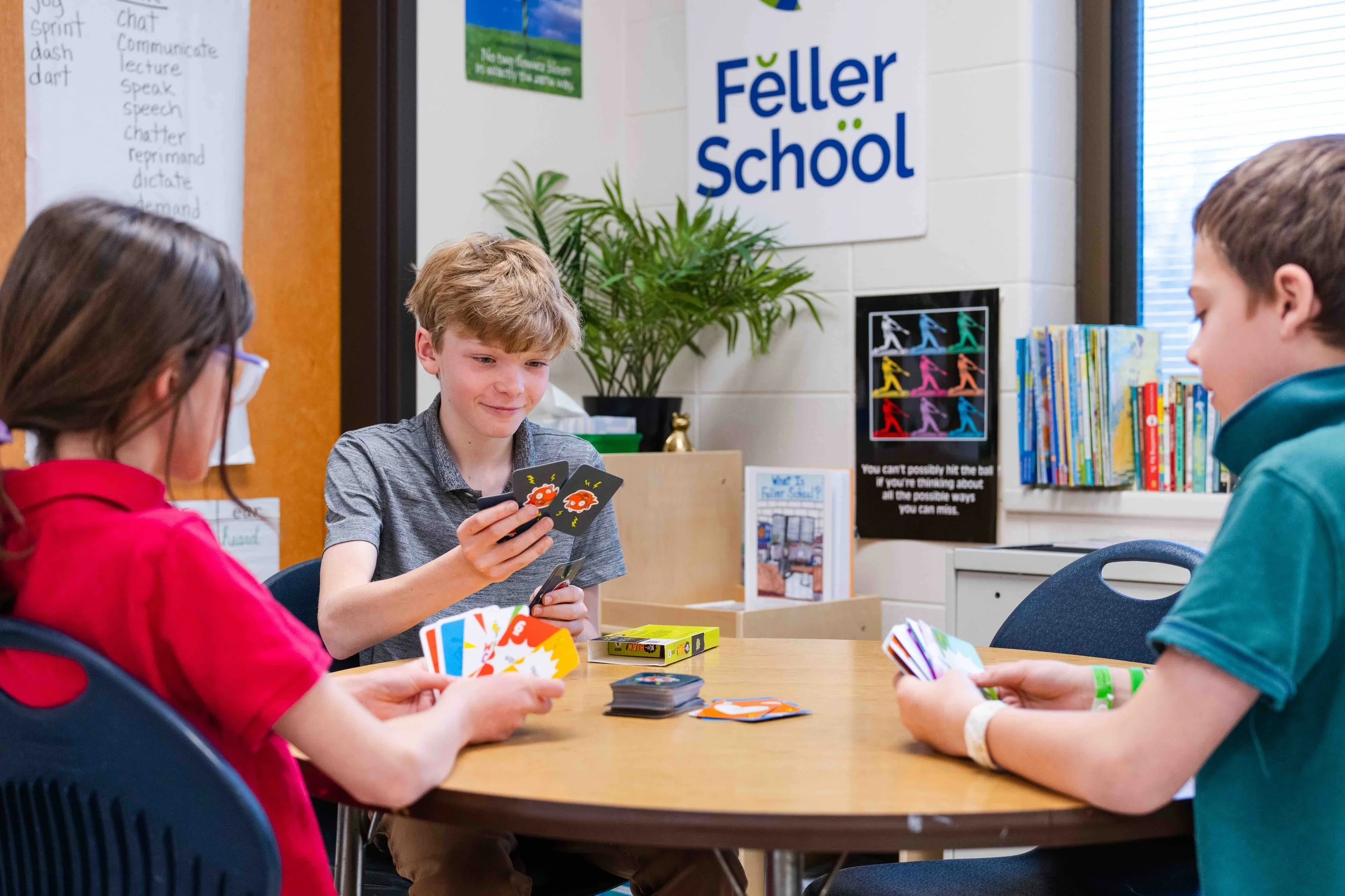 Three children playing a card game at a round table in a classroom with a "Feller School" sign on the wall, bookshelves, and a colorful poster.