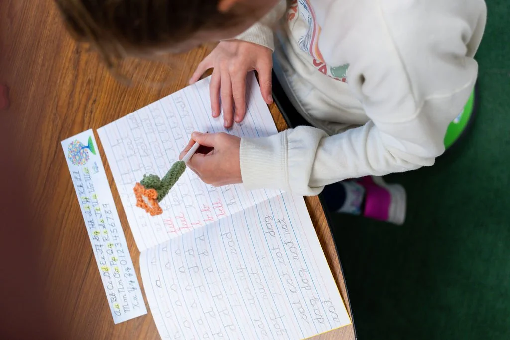 Smiling school children sitting in classroom, boy with brown hair in dark blue jacket and girl with braids in white shirt.
