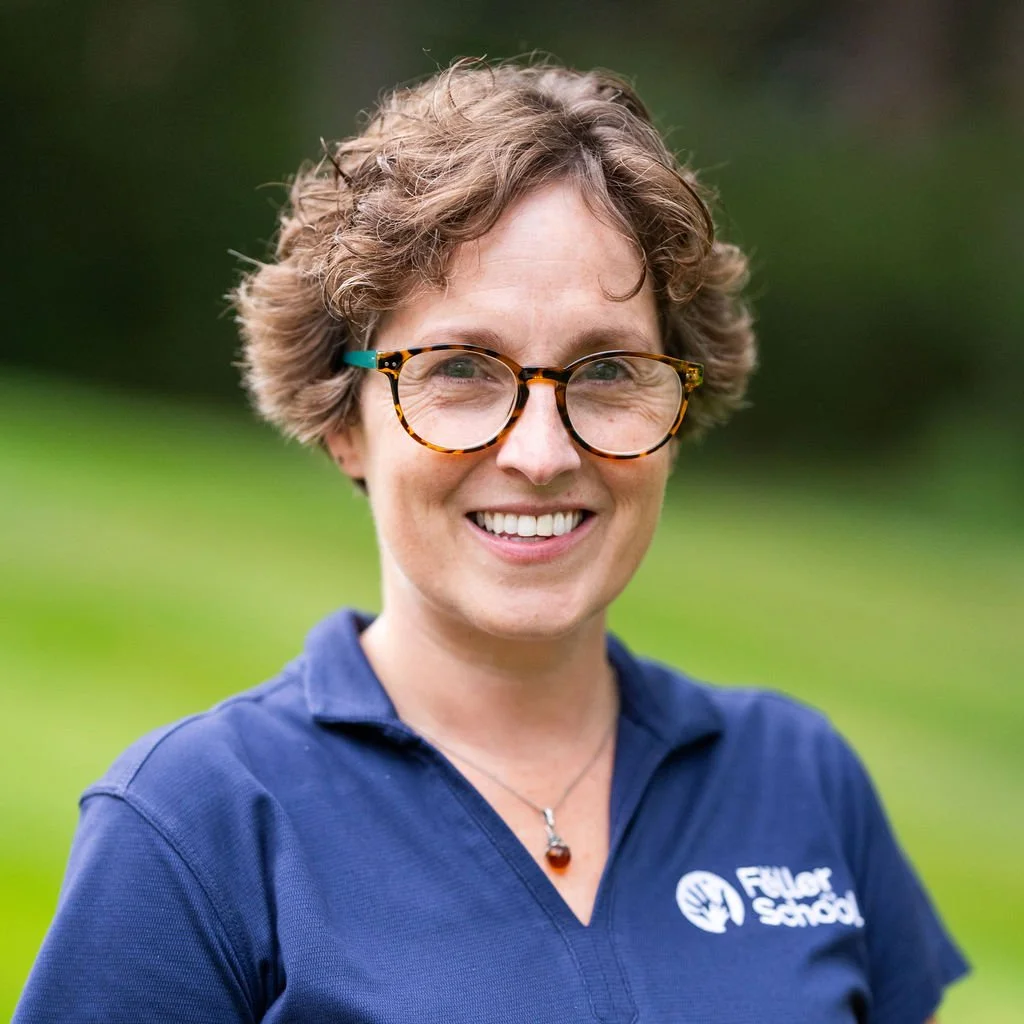 A woman with glasses, short brown hair smiling outdoors, with trees and greenery in the background.