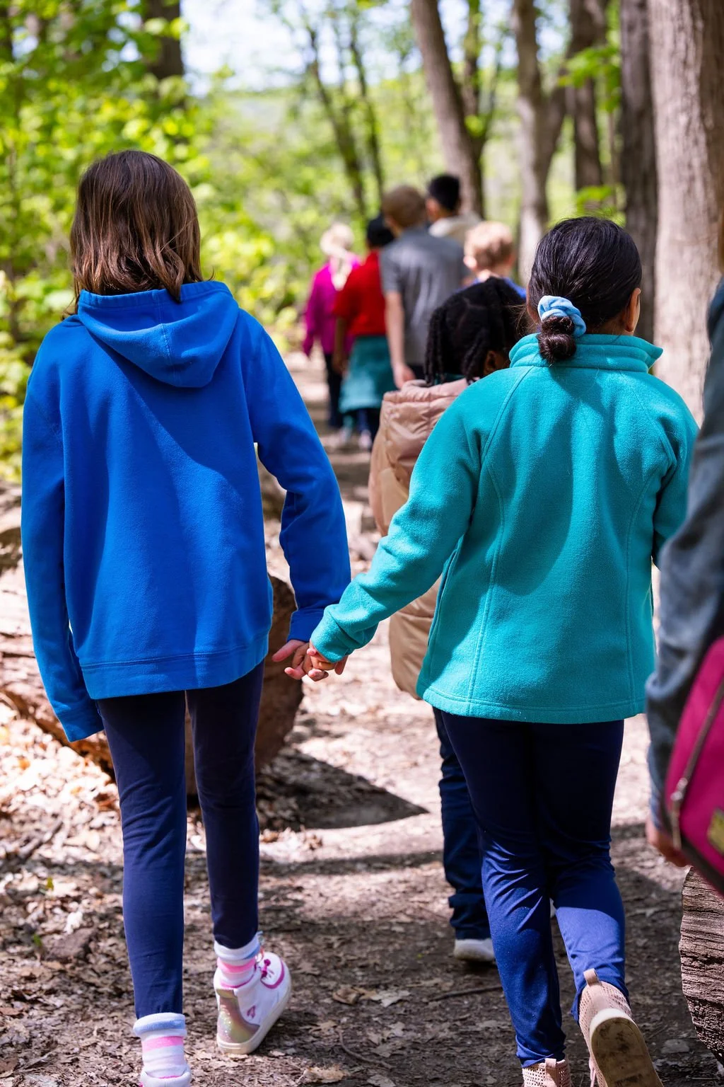 Two children holding hands while walking on a forest trail with other people in the background, surrounded by green trees.