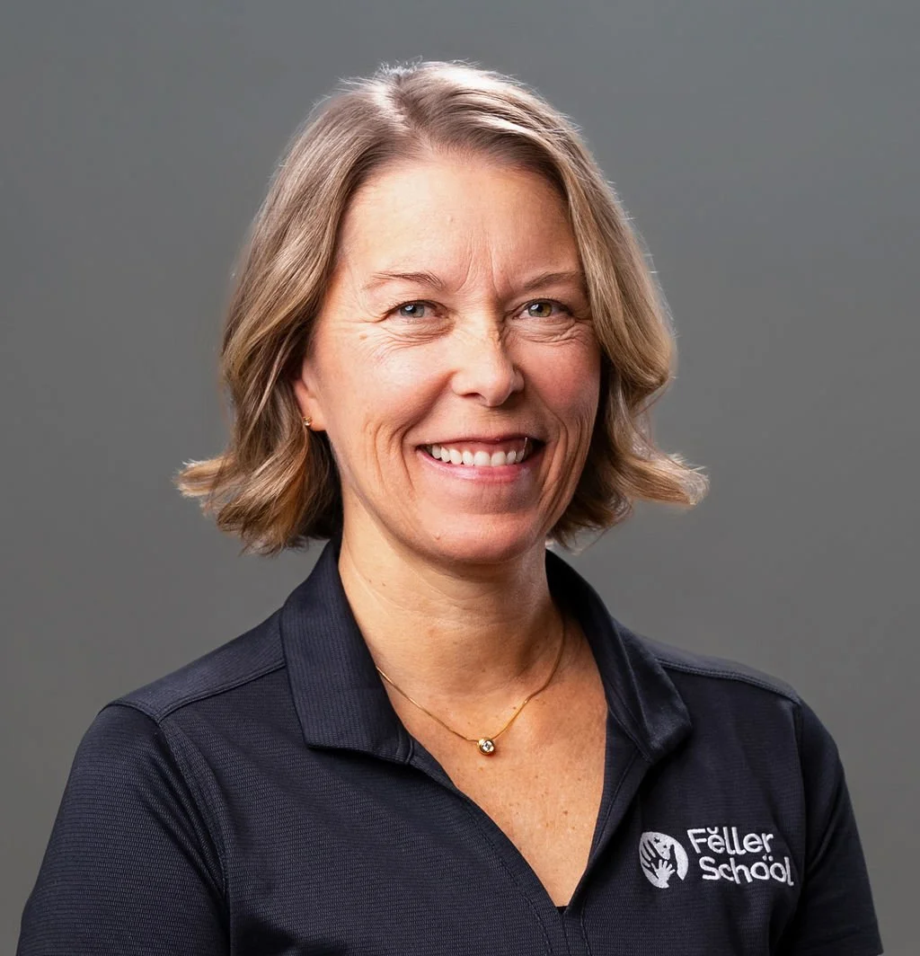 A smiling woman with shoulder-length blonde hair, wearing a white collared shirt and a silver necklace, standing outdoors in front of green leafy foliage.