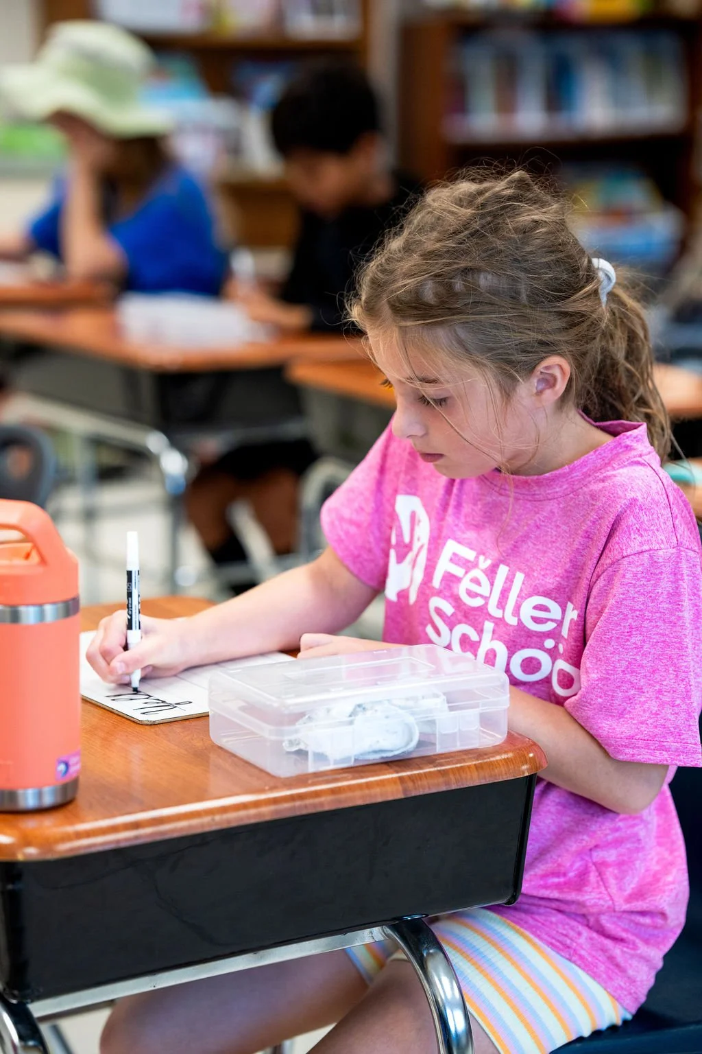 A young girl in a pink 'Feller School' T-shirt writing on a whiteboard with a black marker, sitting at a wooden desk in a classroom.