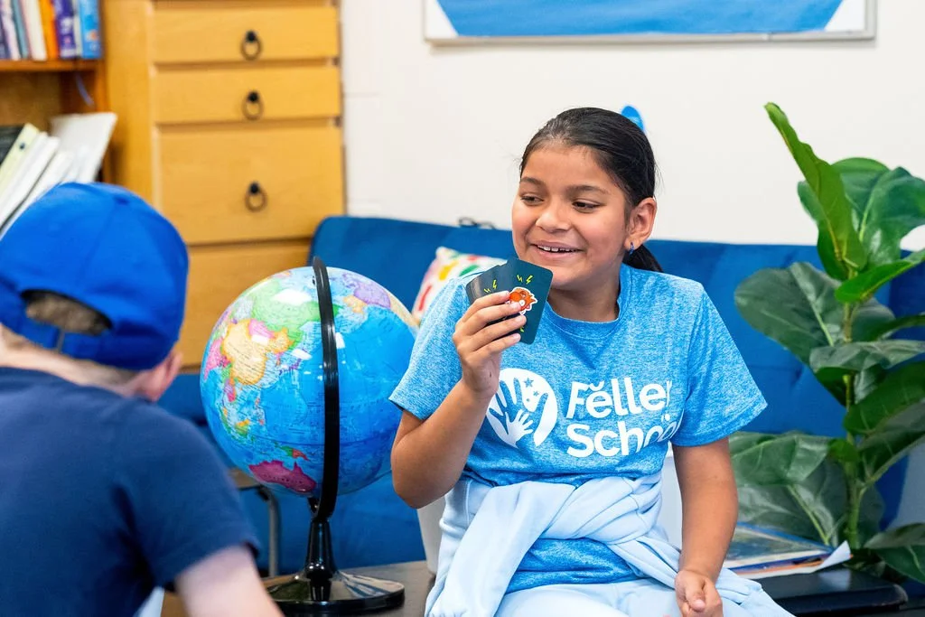 A girl smiling and playing a card game with a person wearing a blue cap in a classroom setting, with a globe, bookcase, and large plant in the background.