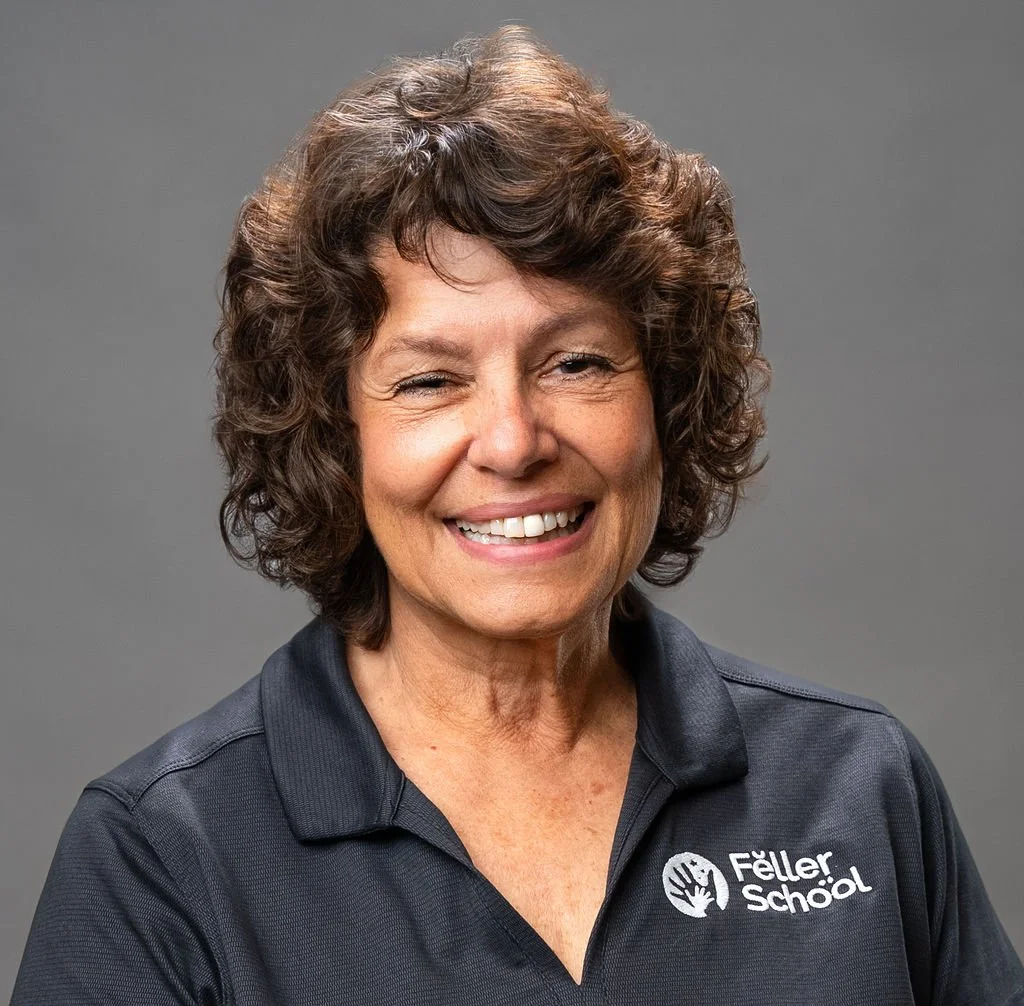 A smiling older woman with short, dark, curly hair standing outdoors in front of a leafy tree.
