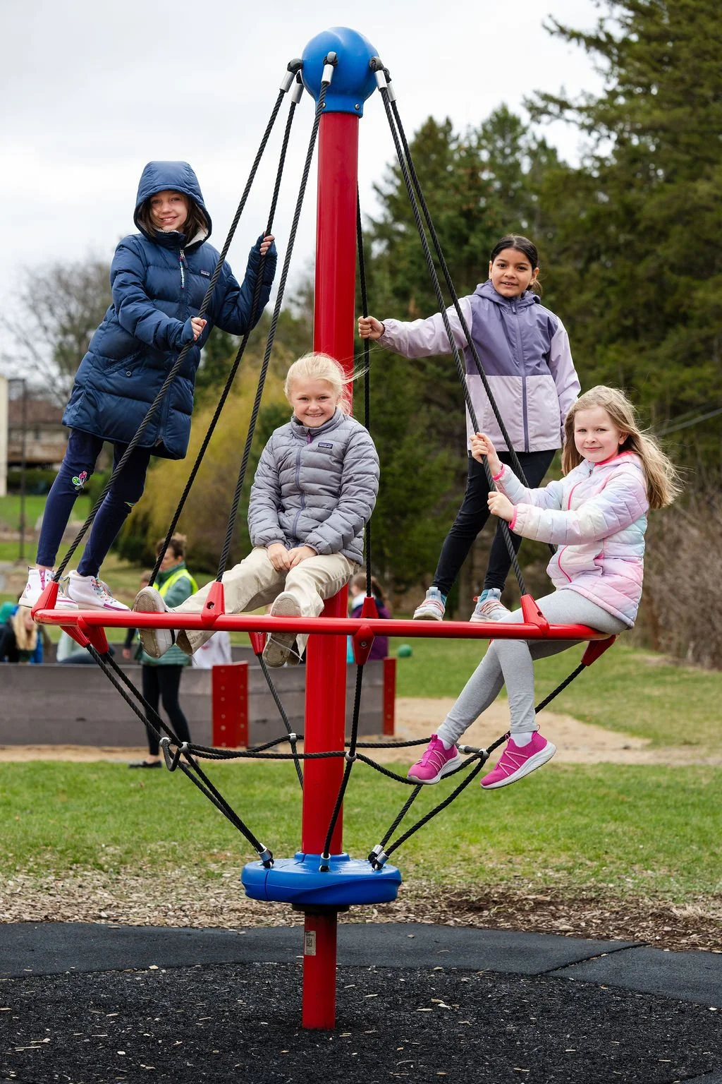 Four girls enjoying a playground on a cloudy day. They are dressed warmly in jackets and are riding a climbing structure with ropes and a platform.