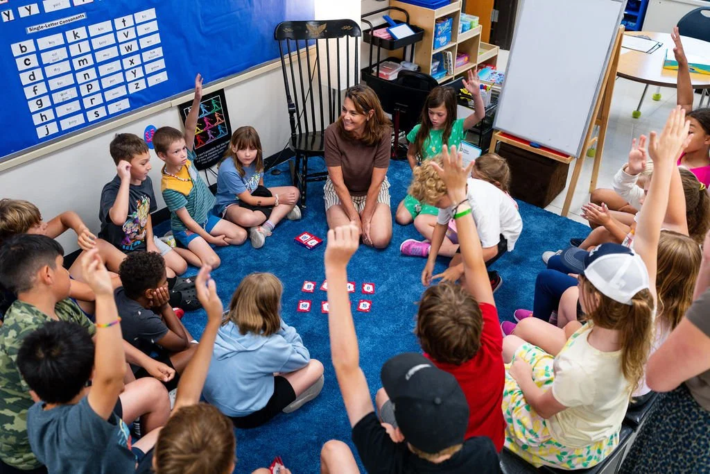 Elementary school classroom with children sitting in a circle on a blue carpet, raising hands, and a teacher in the center. Classroom walls have educational posters, including a letter chart and a whiteboard.