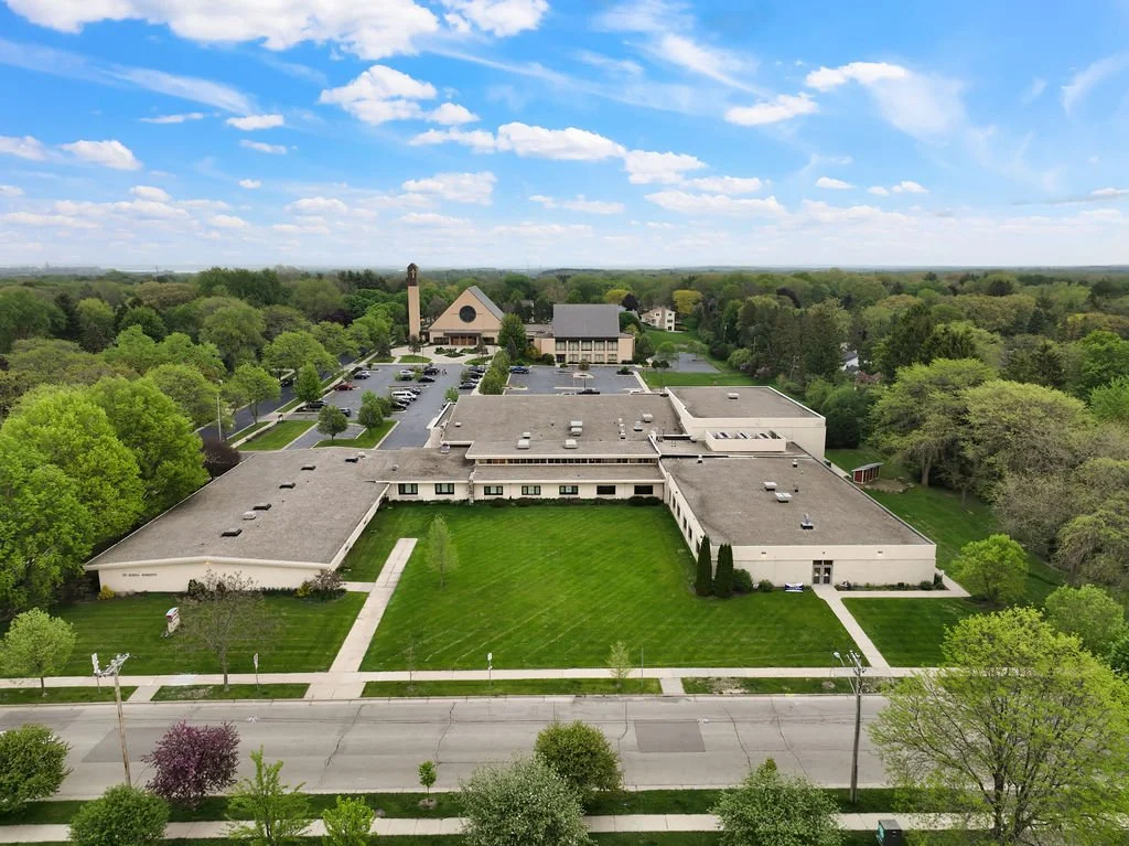 A large building complex with green lawns, surrounded by trees, a parking lot filled with cars, and a street in front, under a partly cloudy sky.