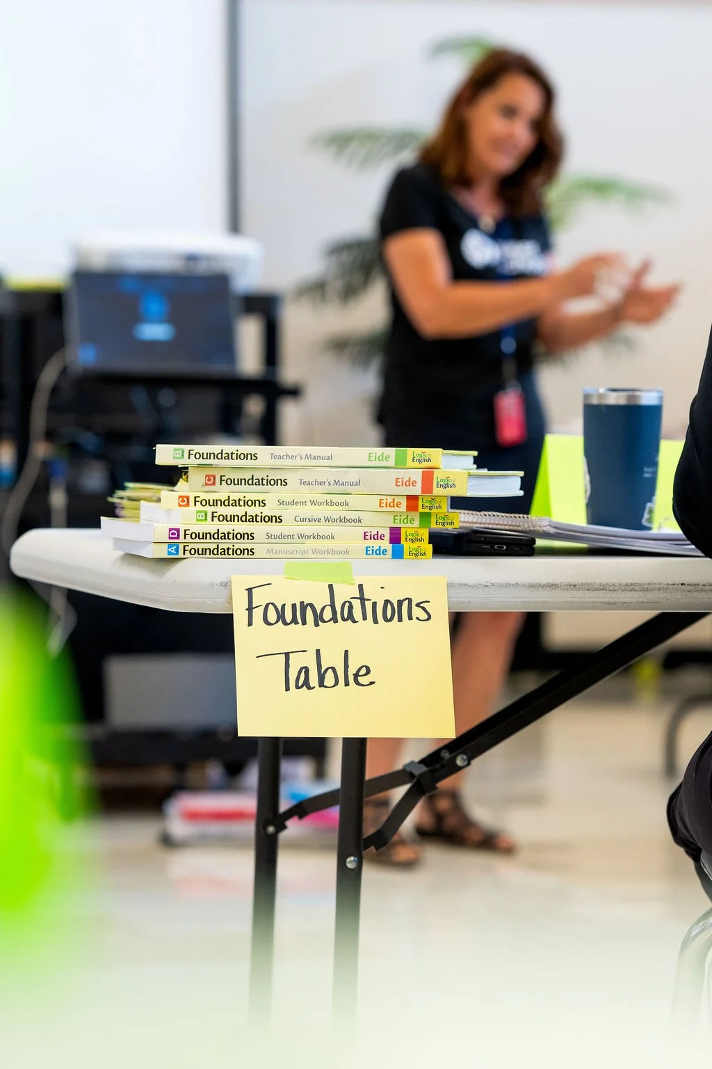 A table with several stacks of educational workbooks and teacher's manuals titled 'Foundations,' labeled 'Foundations Table,' in a classroom or training environment. A woman is blurred in the background, and there is a large water glass and a tablet or notebook on the table.