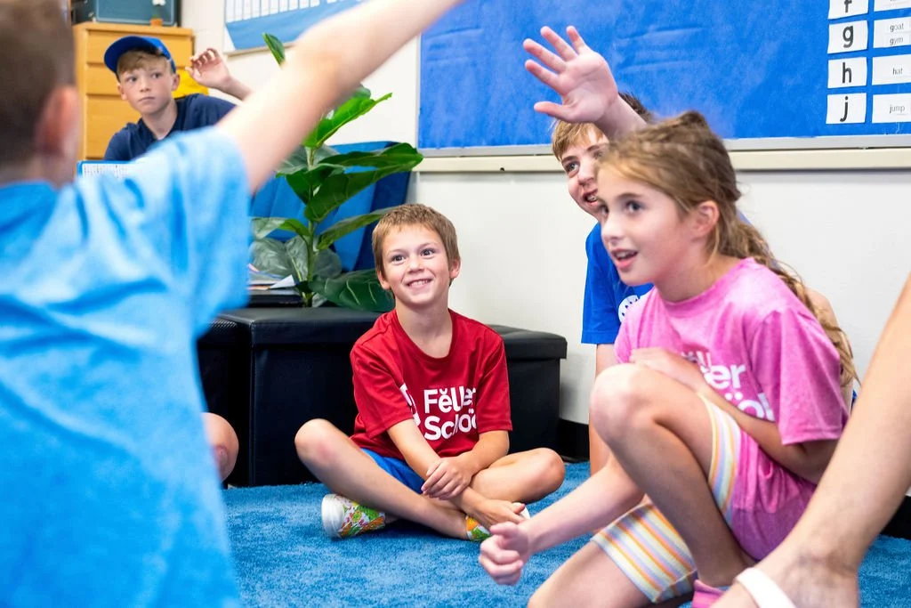 Group of children sitting on blue carpet in classroom engaging in a discussion, with one child raising hand, and a boy in a red shirt smiling.