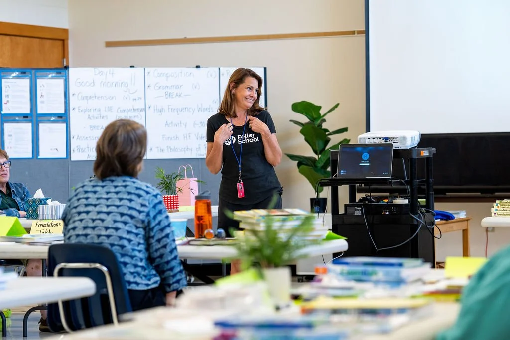 A woman standing in front of a classroom, smiling and gesturing with her hands, with adults seated at tables and whiteboards behind her, in a classroom setting.