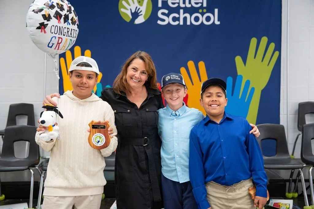 A group of four children and one woman standing together at a graduation event, with a backdrop that reads "Feller School" and a balloon that says "Congrats Grad." The children are holding plush toys and a medal.
