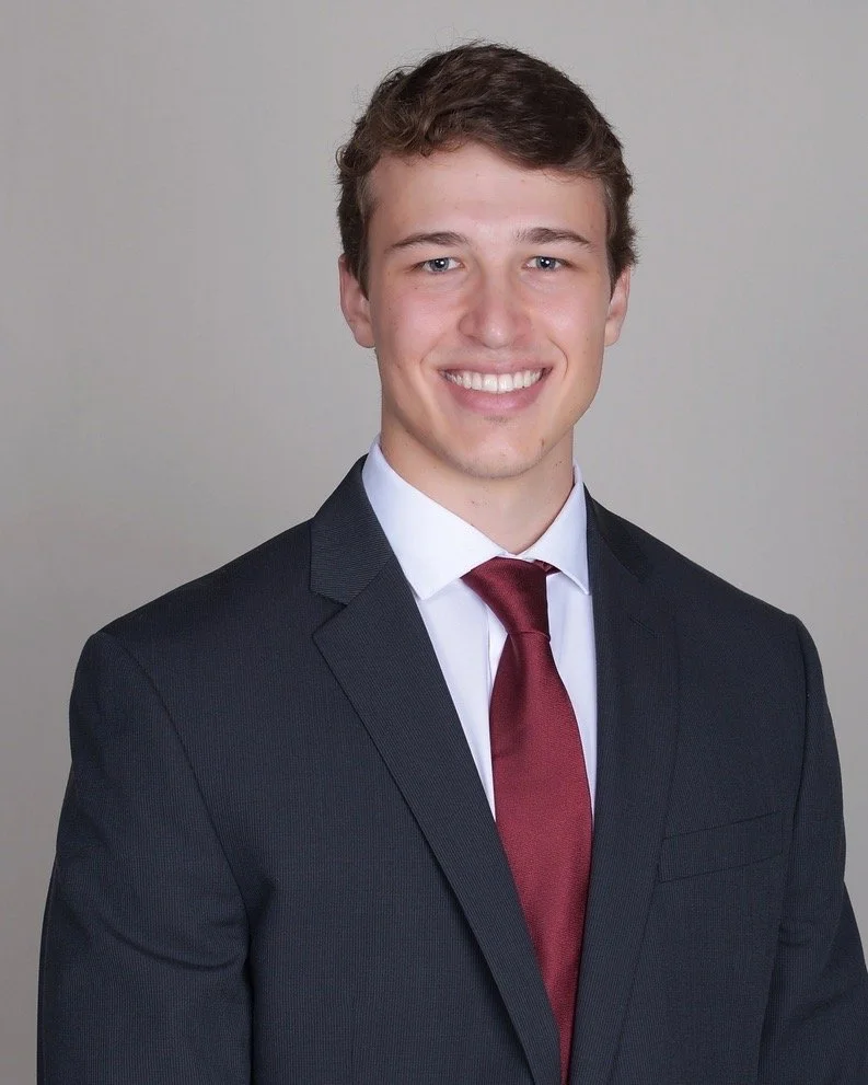 Young man in a dark suit, white shirt, and red tie smiling in a professional portrait against a plain background.