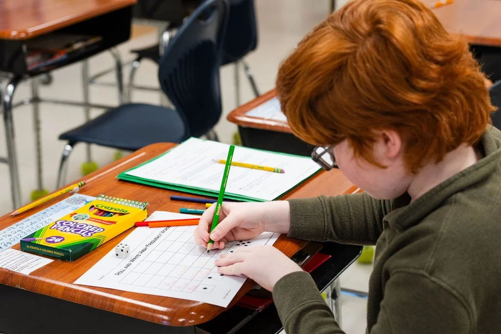 A student with red hair and glasses working at a desk in a classroom, writing on a worksheet with a green pencil. The desk has colored pencils, a die, notebooks, and an open textbook.