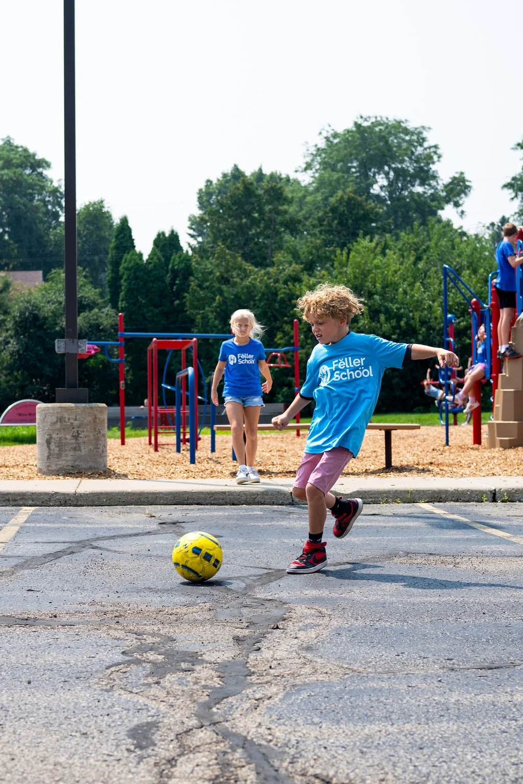 Children playing soccer outside on a school playground, with some children on the climbing structure in the background and others on the pavement