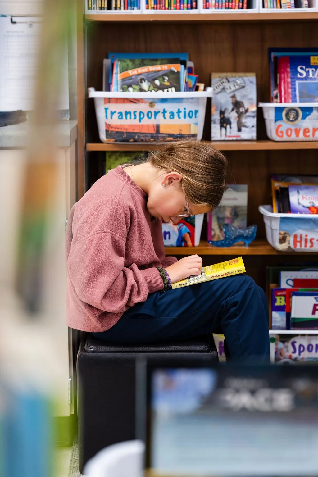 A girl in a pink sweatshirt sitting on a black stool in a library, reading a yellow book. Behind her are shelves labeled 'Transportator,' 'Government,' and 'History' with various books and magazines.