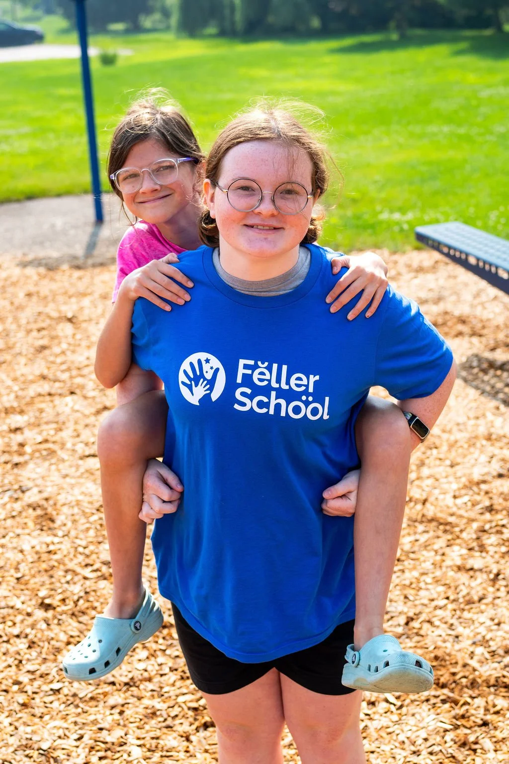 Two smiling girls, one wearing a blue T-shirt with 'Feller School' printed on it, and the other in pink, at a playground with green grass and wood chips.