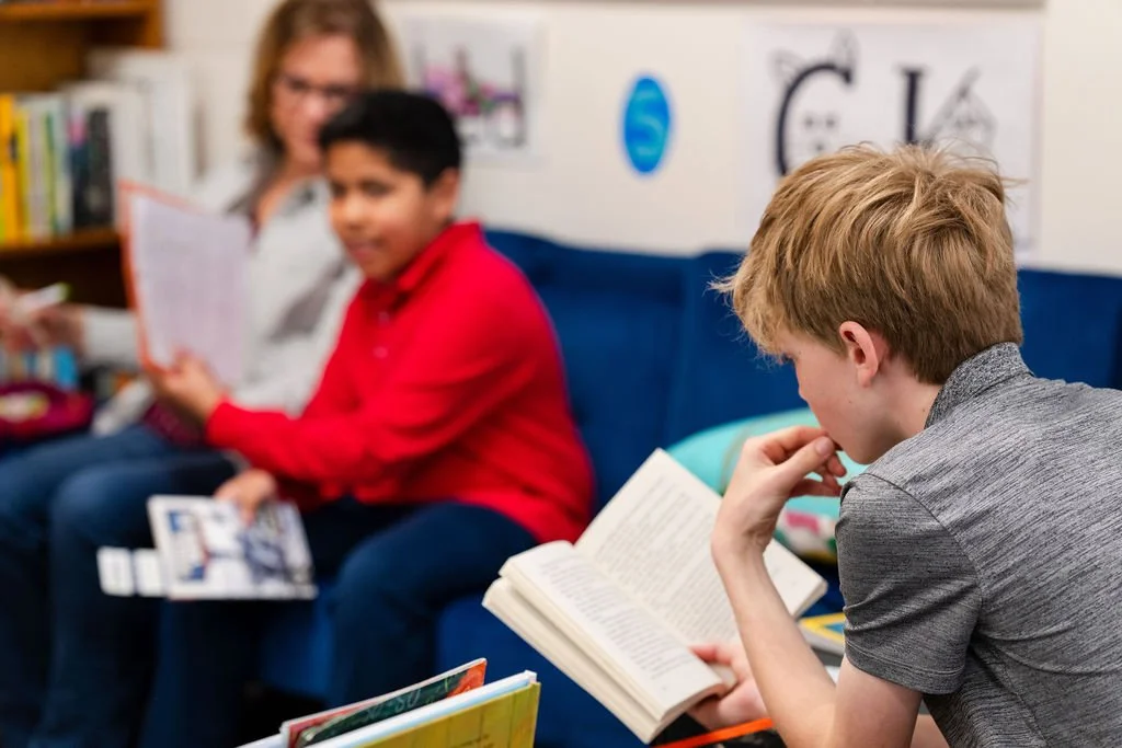 Three children sitting on a blue couch in a library, each reading books with a woman, possibly a librarian, behind them.