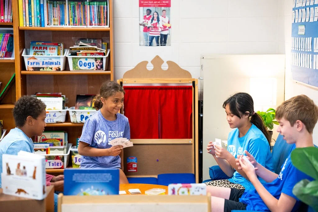 Four children playing a card game in a classroom surrounded by bookshelves and educational posters.