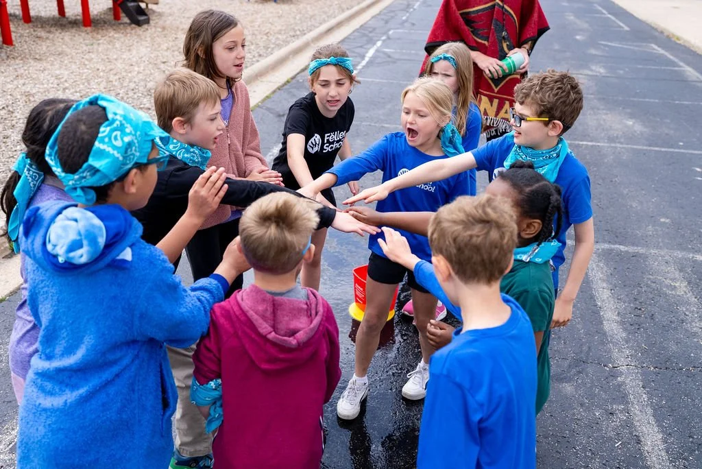 Children participating in a team activity, reaching into a red bucket outdoors on a wet surface.