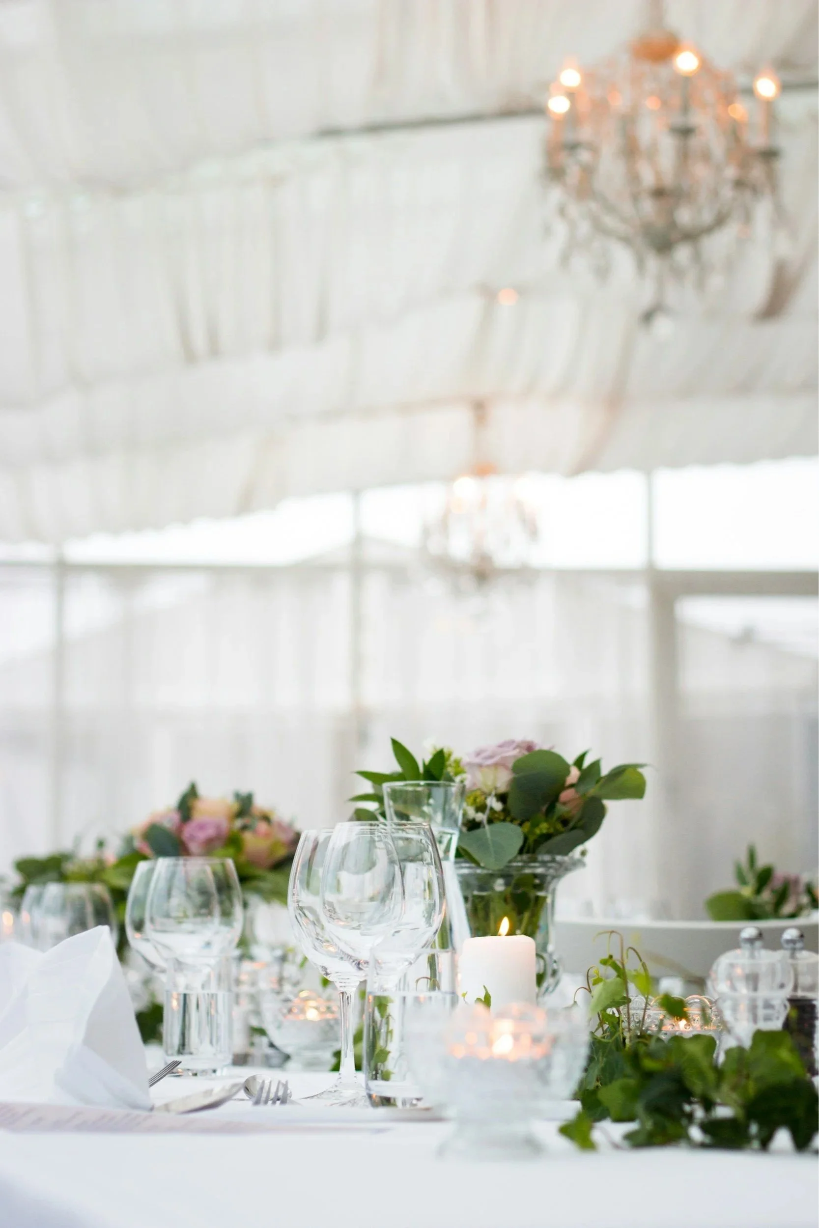Elegant table setting with glasses, candles, floral arrangements, and greenery in a bright, airy venue.