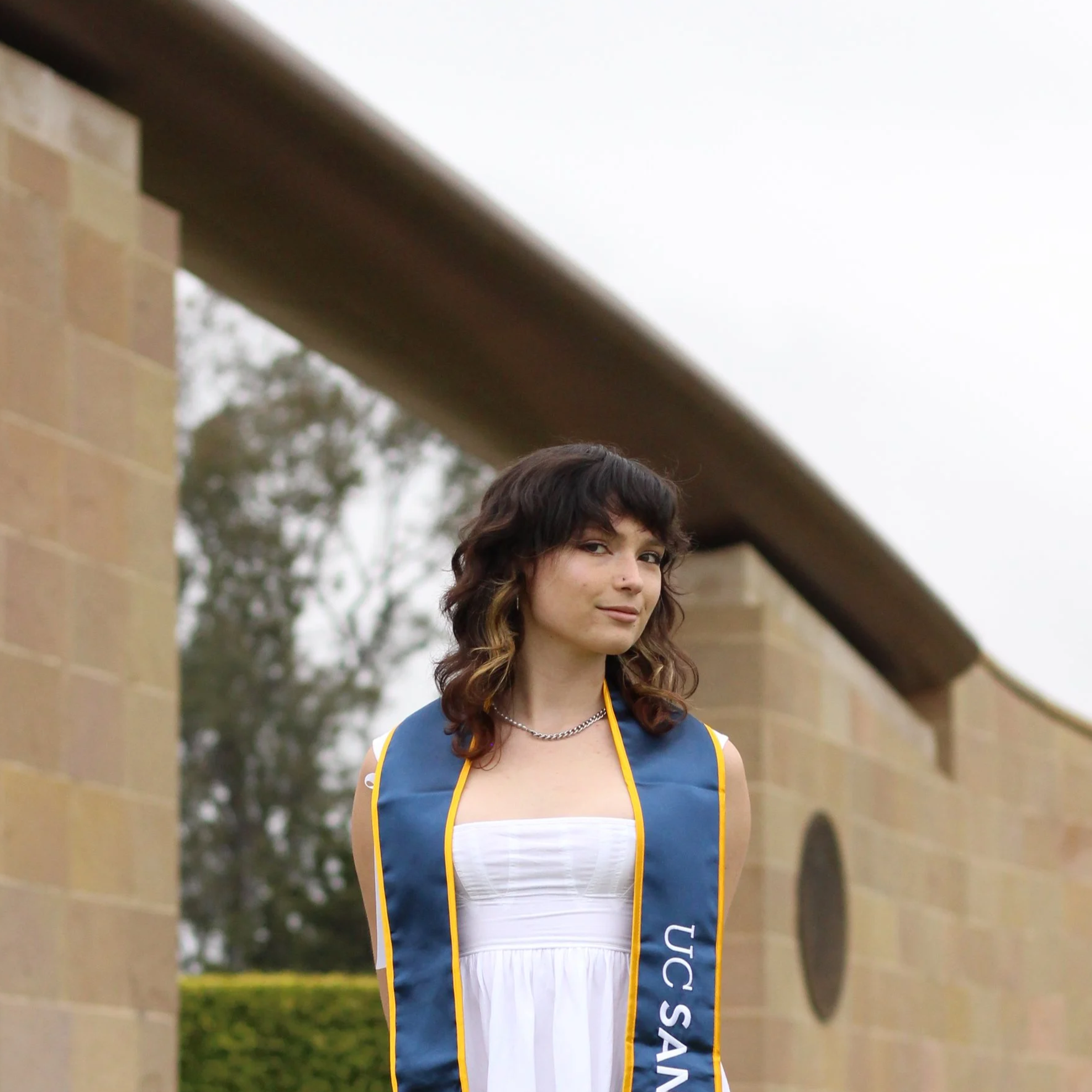 Young woman in a white dress with graduation sash, sitting on a stone ledge outside a university building.