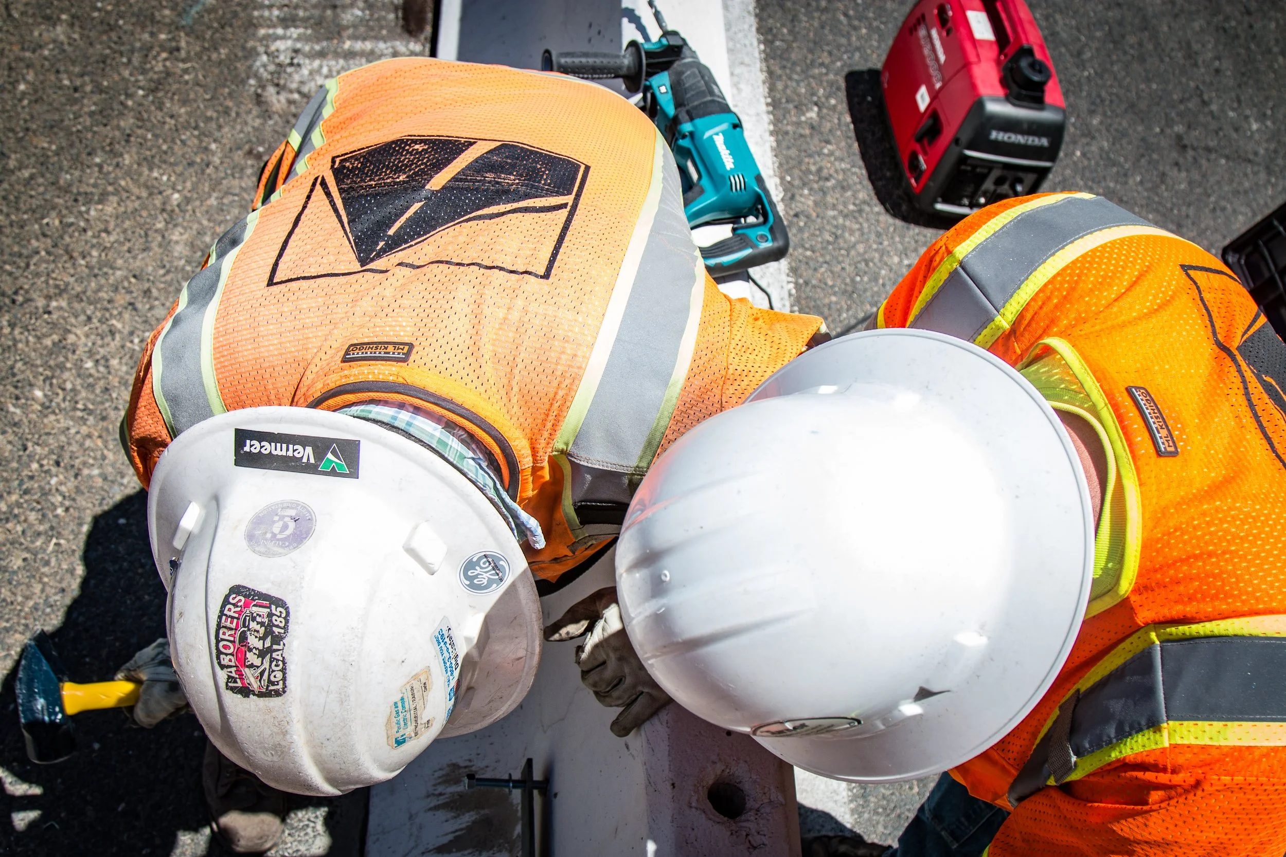 Two construction workers wearing white hard hats and orange safety vests working together on a concrete surface at a construction site.