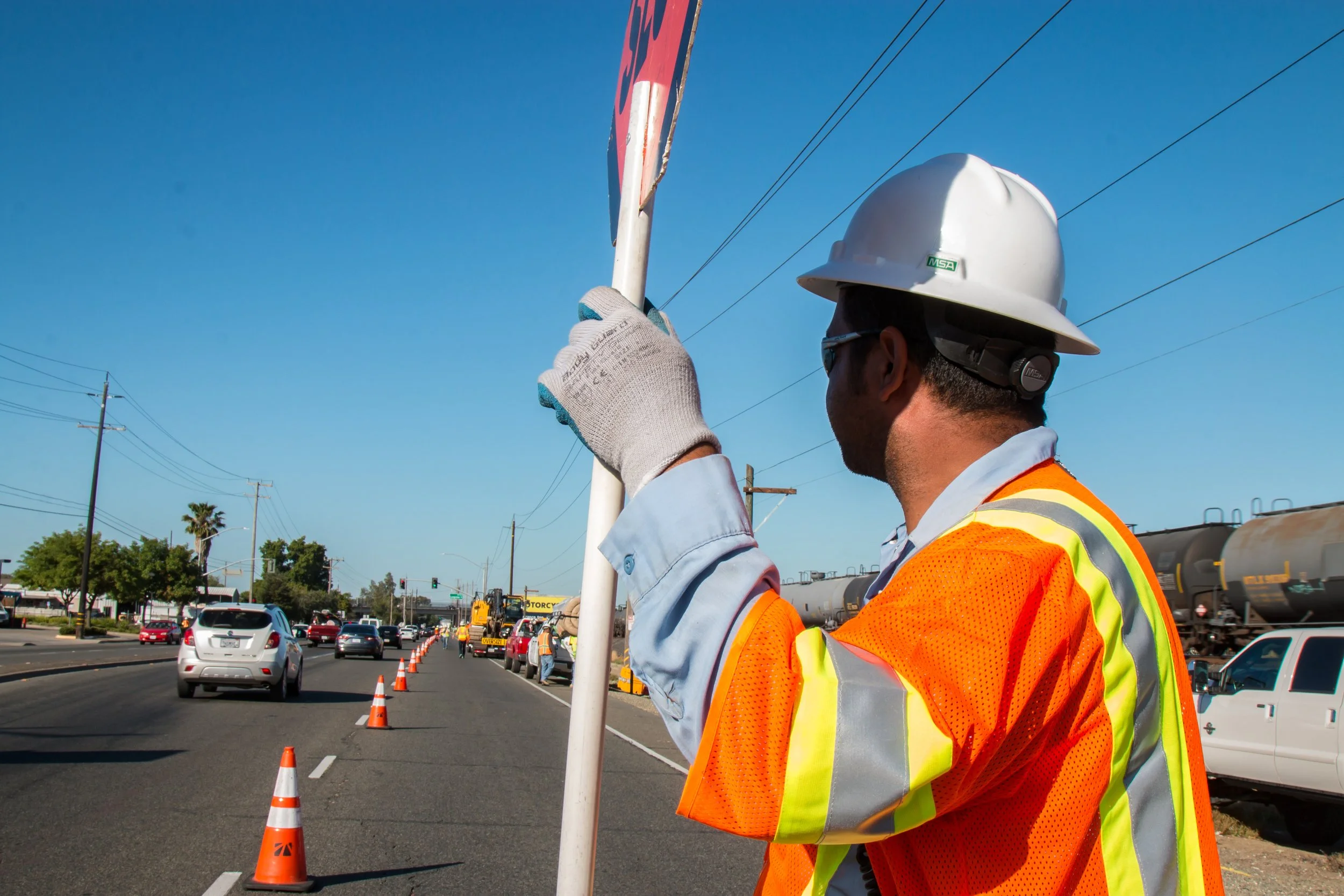 A construction worker wearing a white hard hat, glasses, gloves, and an orange safety vest is holding a stop or traffic sign on a busy street. Cones line the road, with traffic and construction vehicles in the background.